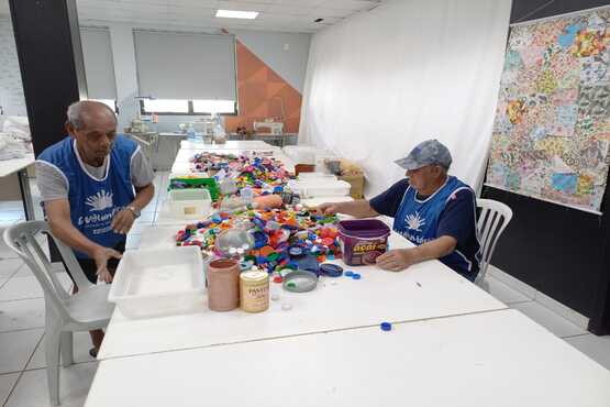 Idosos trabalham em mesa coberta por tampinhas plásticas, para reciclagem. Em troca, entidade recebe artigos esportivos doados para crianças em programas de acolhimento do norte/Nordeste do Estado