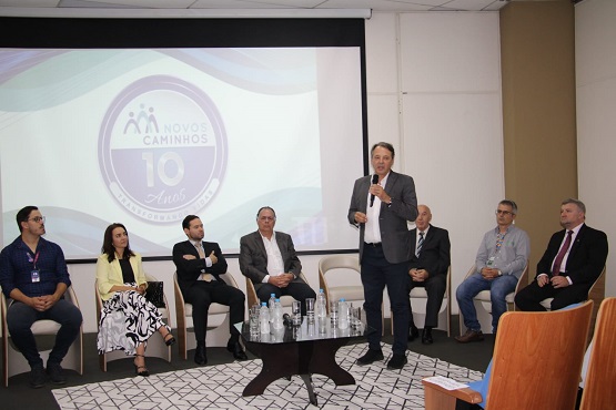 De pé ao centro está o palestrante e sentados ao redor estão os demais membros da mesa de honra