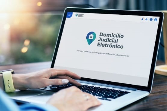 Mão de mulher com as unhas pintadas, usando relógio amarelo no pulso, teclando em notebook que na tela exibe o site do Domicílio Judicial Eletrônico