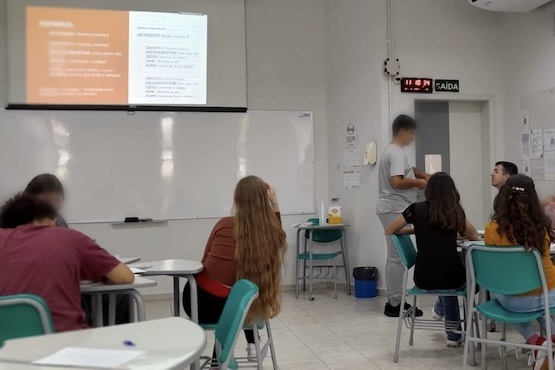 Sala de aula com jovens olhando para slides projetados acima de lousa e conversando entre si