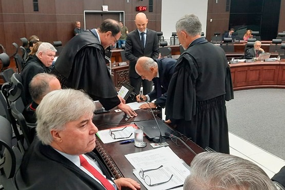Fotografia de homens de toga no pleno, assinando documento em cima de mesa de bancada de madeira