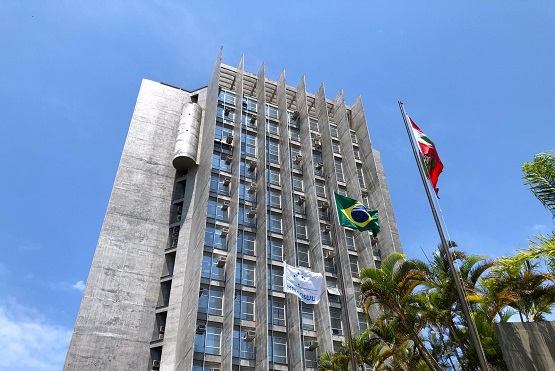 Fotografia de um prédio de doze andares feita de baixo. Ao lado é possível ver três bandeiras e o topo de palmeiras. Ao fundo, o céu azul.