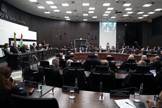 Homens e mulheres sentados em bancada semicircular de madeira voltados para mesa de autoridades em sessão do Tribunal de Justiça.