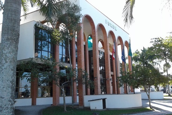 Visto de diagonal, prédio branco de dois andares com colunas e arcos na fachada. Árvores e bandeiras na frente do edifício.