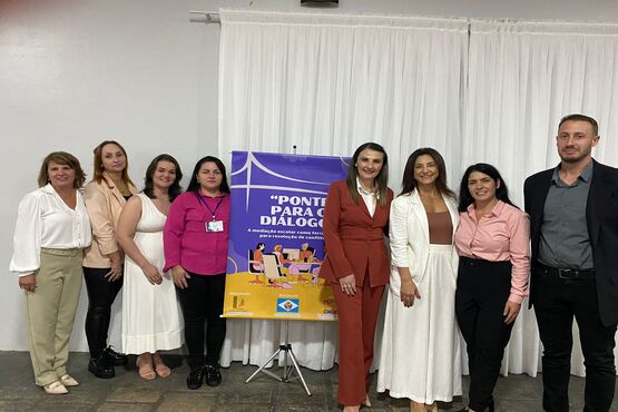Ao centro da foto está o banner do programa "Ponte para o Diálogo" nas cores roxa, amarela e branca. Ao lado esquerdo do banner estão quatro mulheres e, ao direito, três mulheres e um homem, todos posam para a fotografia.