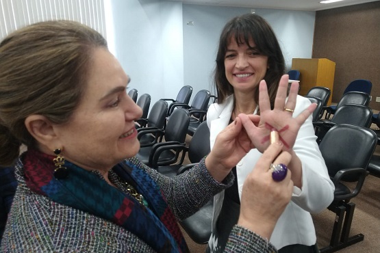 Mulher mais velha desenhando um X vermelho na palma da mão de mulher mais nova com uma caneta. Ambas sorriem e observam o desenho sendo feito.