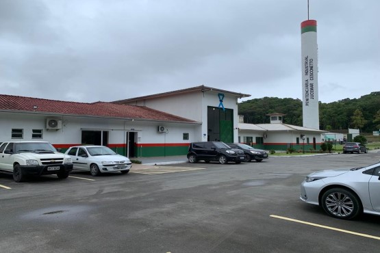 Construção de um andar branca, com faixas verdes e vermelhas na parte inferior. Ao lado uma torre circular nas mesmas cores. Em frente, um estacionamento com carros.