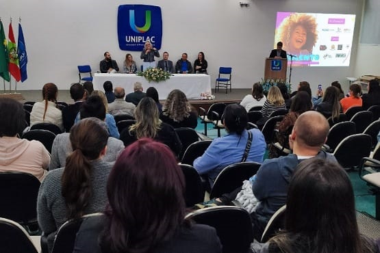 Fotografia de pessoas assistindo palestra. À frente, mesa retangular com toalha branca e flores. Atrás dela, os palestrantes. Na parece a logo da Uniplac.