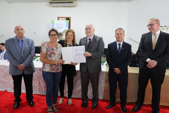 Fotografia de pessoas lado a lado, no centro, um homem entrega o título de propriedade para uma mulher.