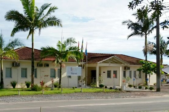 Fachada de edificação térrea. Na frente da construção tem uma placa escrito “FÓRUM” e plantas de médio e grande porte.