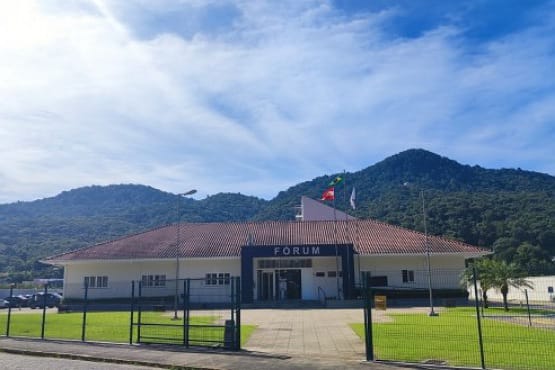 Fachada de edificação térrea rodeada por grama e cerca. Em cima da porta de entrada da construção está escrito “FÓRUM” e ao lado estão hasteadas três bandeiras.