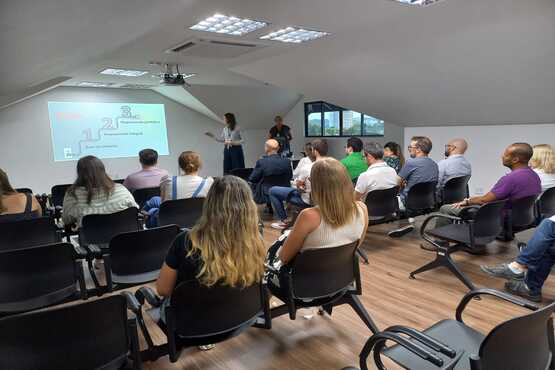 Fotografia de pessoas assistindo uma palestra. Na frente da sala tem uma parede branca com slides sendo projetados e uma mulher de saia e camiseta branca os apresenta.