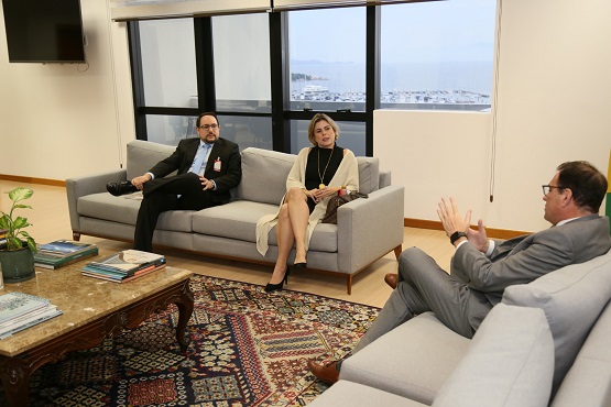 Três pessoas sentadas em sofás conversam. No sofá à esquerda, um homem e uma mulher olham em direção a outro homem, que está sentado em um sofá à direita, de costas para foto e gesticulando.