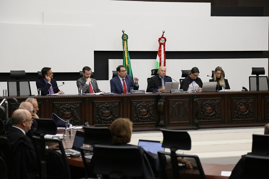 Ao centro da imagem, homens e mulheres sentados atrás de uma bancada, que é utilizada para apoiar papeladas, copos de água, microfones e computadores. Ao fundo, dois mastros e bandeiras. No primeiro plano da imagem, que está desfocado, estão homens e mulher sentados de costas para a foto.