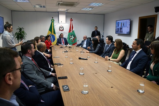 Homens e mulheres sentados à mesa. Ao fundo estão duas bandeiras e um banner do Tribunal de Justiça do Estado de Santa Catarina.