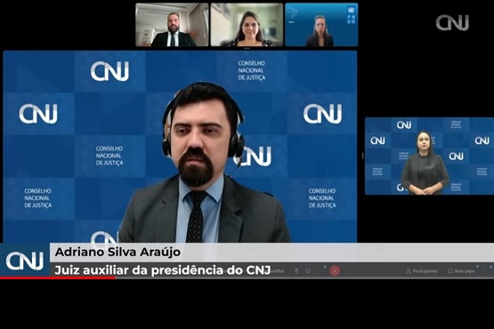 Imagem do webinário com o juiz auxiliar da presidência do CNJ, Adriano Silva Araújo, ao centro e em fundo azul composto por quadrados menores escrito CNJ. Tela dos demais participantes na parte superior da imagem. Na lateral direita está a intérprete de libras.