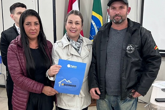 Três pessoas posam para a foto. Da esquerda para direita estão duas mulheres seguram uma pasta azul, que possui na capa um contorno em branco de uma casa e a frase “#SONHO” nas cores laranja e azul escuro. e um homem. Ao fundo estão três bandeiras.