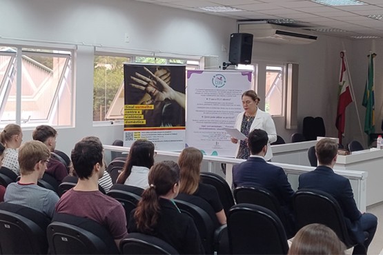 Pessoas sentadas de costas para foto olham para uma mulher, que está de pé ao centro da imagem e virada em direção daqueles que estão sentados, lendo um papel. Ao lado direito, estão dois banners explicativos sobre violência doméstica e o programa CEAV nas Comarcas.