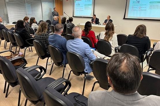 Pessoas sentadas de costas para foto olham para dois homens sentados em uma mesa, que estão discutindo sobre temas relacionados a Governança e Meio Ambiente.