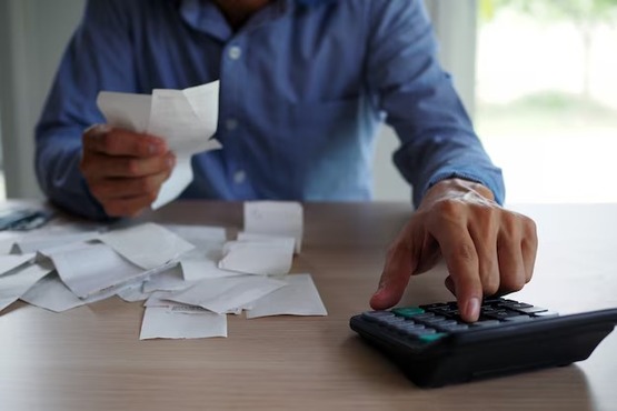 Com uma das mãos, uma pessoa clica em tecla de calculadora e , na outra, segura papéis em formatos retangulares, que estão também espalhados por cima da mesa de madeira.