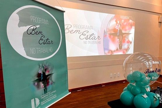 Ao lado esquerdo da foto está situado na cor verde-azulado um banner do Programa Bem-Estar no Trabalho. Ao fundo uma projeção em uma parede branca com o nome do programa e a frente uns balões na mesma cor do banner do programa.