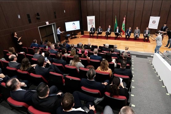 Um grupo de pessoas está sentada nas cadeiras vermelhas do Auditório Pleno do TJ, assistindo os 7 homens que estão sentados no palco em suas frentes. Fotógrafos tiram fotos ao redor.