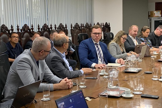 Lateral esquerda de uma mesa comprida, com Homens e mulheres sentados ao redor. Sobre ela copos, celulares, notebooks e papeladas.