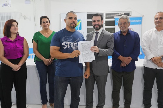 Foto de seis pessoas em pé, sendo quatro homens e duas mulheres. Dois dos homens estão segurando uma folha de papel com um texto.
