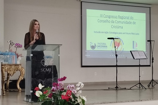 Ao lado esquerdo da fotografia, mulher atrás de púlpito fala em microfone e, no lado direito, está projetado na parede “III Congresso Regional do Conselho da Comunidade de Criciúma” em letras maiores, logo abaixo está escrito “Inclusão em Ação: Estratégicas para um Futuro Mais Justo” e, por fim, as logos dos apoiadores do congresso.
