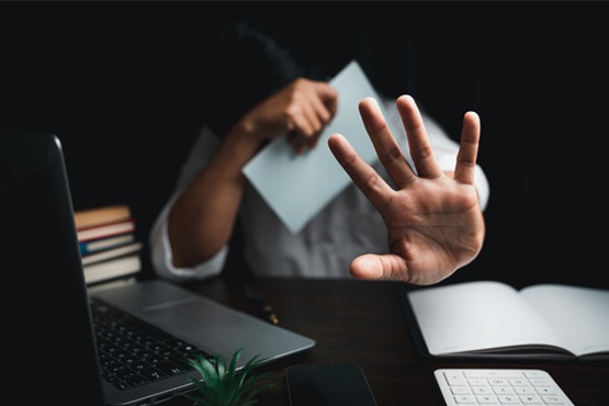 Pessoa cobre rosto com o braço direito e estica a mão esquerda, de um modo que sua palma esteja toda aberta para a foto e em primeiro plano. A ambientação é composta pela pessoa sentada em uma mesa que possui computador, livros e papéis.