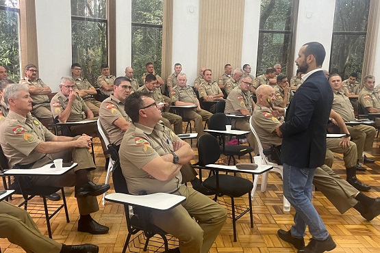 Grupo de Policiais Militares dispostos em cadeiras enfileiradas dentro de uma sala. Um homem em pé e de terno, dando aula do cursos para o aprimoramento profissional e segurança institucional.