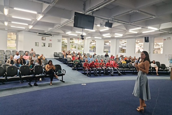 Grupo de pessoas sentadas em cadeiras enfileiradas e uma mulher em pé.
