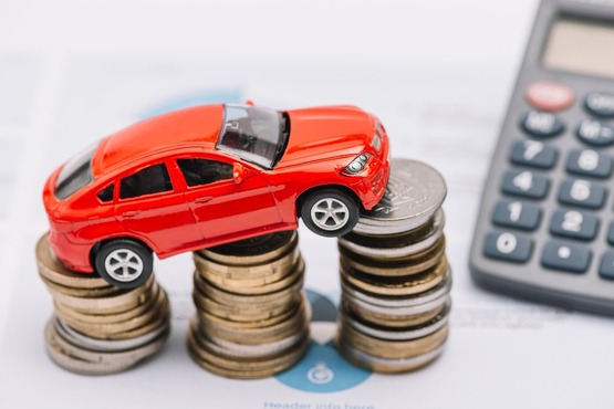 Carrinho de brinquedo colocado sobre três pilhas de moedas, e ao lado uma parte de uma calculadora.
