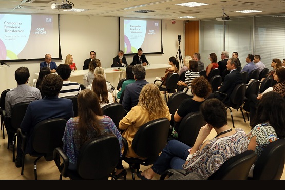 Foto de um grupo de pessoas de costas em uma sala, sentadas em cadeiras enfileiradas de frente com outras pessoas também sentadas em cadeiras e uma mesa entre eles.