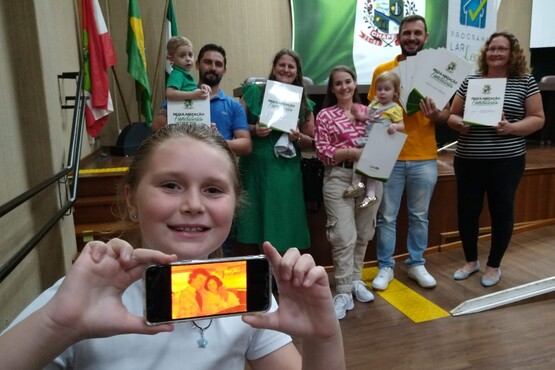 Dois homens, três mulheres e três crianças posam para uma foto, uma das crianças está em primeiro plano, mostrando um celular com uma foto antiga de um homem e uma mulher.