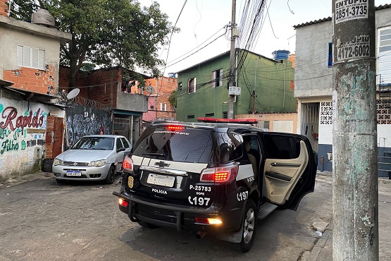 Viatura da policia civil esta em um bairro com algumas casas, o carro é preto e está com uma das portas traseiras aberta.