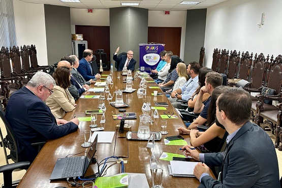 Grupo de pessoas dentro de uma sala de reunião, sentadas em cadeiras ao redor de uma mesa comprida. Sob a mesa estão alguns copos de águas, papéis  e alguns pertences pessoais.