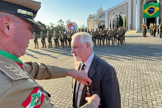 A imagem mostra uma cerimônia militar ao ar livre. No centro da imagem, há um oficial militar, usando uniforme bege com insígnias no ombro e um chapéu, colocando uma medalha ou insígnia no paletó de um homem mais velho de cabelos brancos, que está vestindo um terno escuro e gravata. Ao fundo, há uma formação de soldados em uniforme, e uma grande bandeira do Brasil está visível. Também é possível ver um arco ou portal branco ao fundo, que parece ser a entrada de um quartel ou instalação militar.