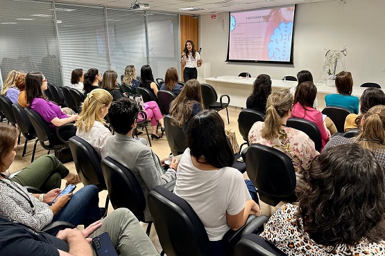 Um grupo de pessoas de costas para a fotografia olham para uma mulher que está palestrando na frente da sala.