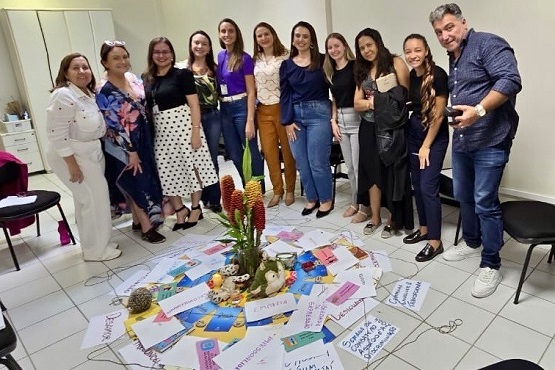 Dez mulheres e um homem posam para foto. No chão, à frente deles, estão espalhados em um círculo alguns papéis, pelúcias e um vaso de planta.