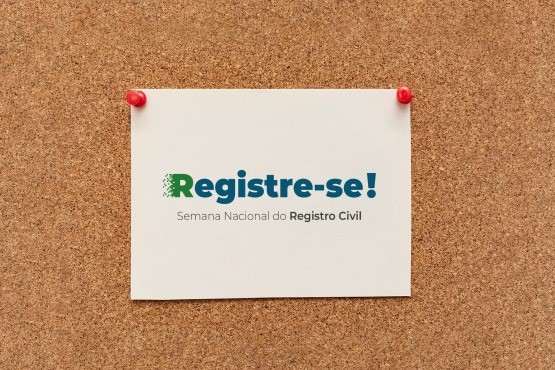 Arte gráfica da 2ª Semana Nacional do Registro Civil - “Registre-se”. No meio de um quadro de cortiça está uma folha com a palavra "registre-se” em azul, sendo a primeira letra em maiúscula e na cor verde, e, em baixo e em uma fonte menor, a frase “Semana Nacional do Registro Civil”. O papel é preso por alfinetes vermelhos nas laterais superiores.