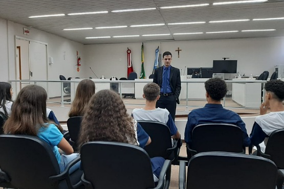 Pessoas sentadas de costas para foto no espaço em que ocorrem as sessões do Tribunal do Júri na comarca de Videira olham para um homem engravatado que está de pé na frente do grupo.