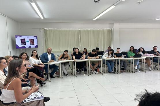 Grupo de pessoas estão em uma sala sentados em cadeiras escolares.