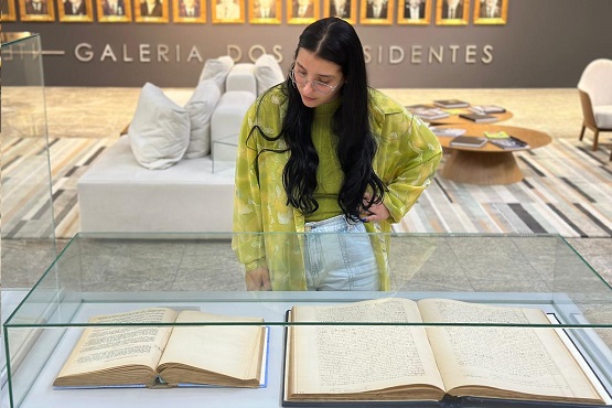 Jovem mulher olha para dois livros abertos que estão expostos em um redoma quadrada de vidro.