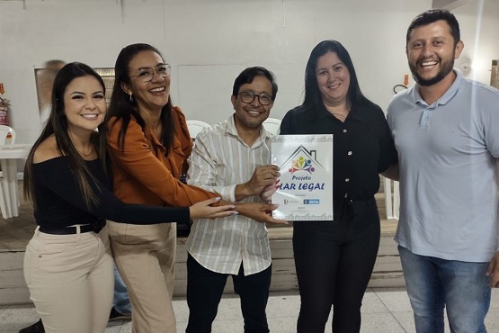 Três mulheres e dois homens posam sorridentes para foto. O homem que está no centro da imagem segura, em conjunto com a mulher do seu lado, uma pasta escrito projeto Lar Legal.