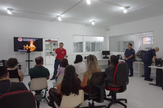 Foto de um grupo de pessoas de costas, em uma sala sentadas em cadeiras enfileiradas, com os olhares direcionados a um homem fardado com a farda do Corpo de Bombeiros Militar de Santa Catarina, ao lado de uma televisão na parte da frente da sala.