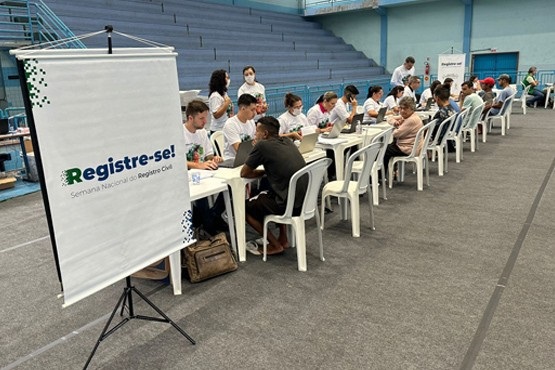 Foto mostrando os atendimentos, que tem o intuito de oportunizar a promoção da cidadania para as populações vulneráveis, feitos em mesas plásticas em um ginásio com paredes azuis. No lado esquerdo das mesas de atendimento está o banner do programa “Registre-se”.
