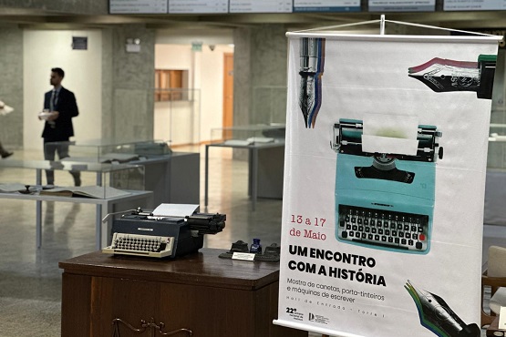No lado direito da foto está o banner da exposição “Um encontro com a História” e, ao fundo, está a exposição desfocada.