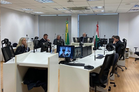 Foto tirada de uma sessão com magistrados sentados e olhando para computadores. Ao fundo estão duas bandeiras.