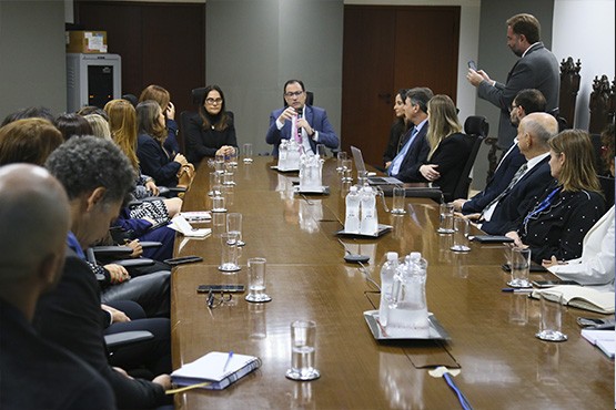 Foto tirada de pessoas sentadas à mesa. Um homem engravatado de óculos, que senta na ponta oposta de quem tirou a fotografia, fala ao microfone.